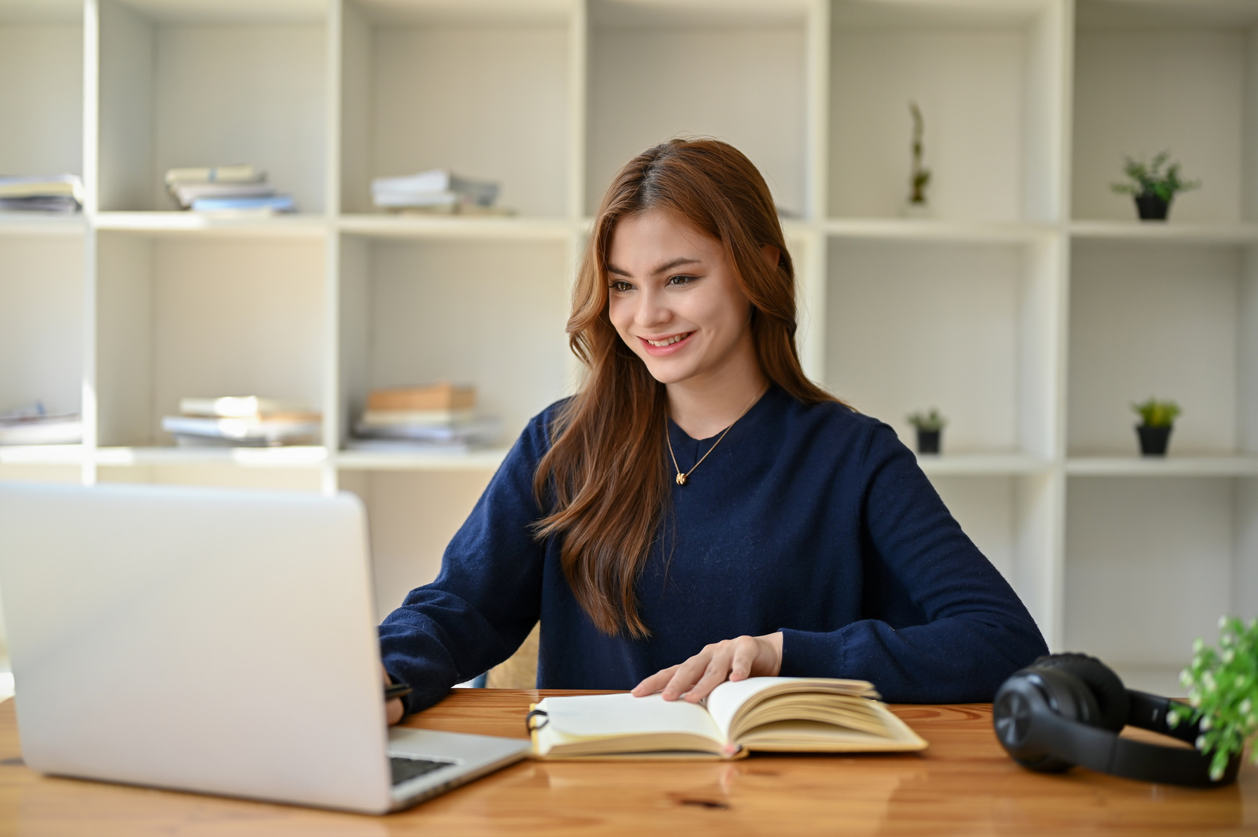 A charming Asian woman in a cozy sweater is working on her assignment on her laptop